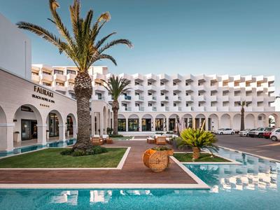 Weißes Hotelgebäude mit Arkaden, Palmen und Wasserbecken im Vordergrund bei klarem Himmel.