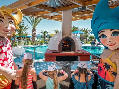 Bambini giocano vicino a una piscina con un forno per pizza all'aperto e maschere colorate.