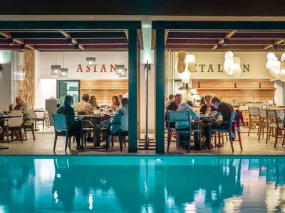 Persone che cenano in due ristoranti adiacenti accanto a una piscina illuminata di notte.