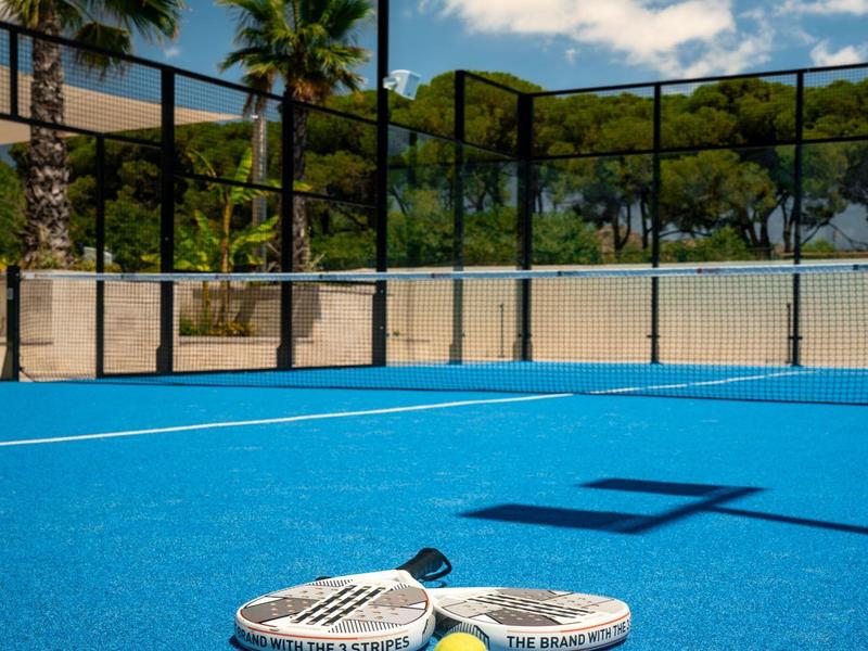 Campo da padel blu con due racchette e una palla, circondato da palme e alberi.