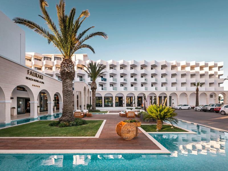 Hotel bianco con piscina, palme e terrazze sotto un cielo sereno.