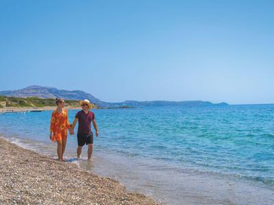 A couple walks barefoot along a pebbled beach, surrounded by blue sky and sea.