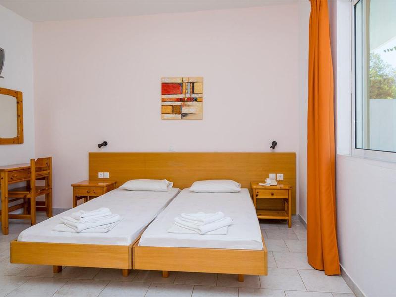 Brightly furnished hotel room with two single beds, desk, and window with orange curtain.