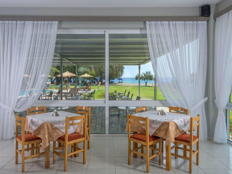 Interior of a restaurant with tables and chairs, overlooking a terrace and garden with sun umbrellas.