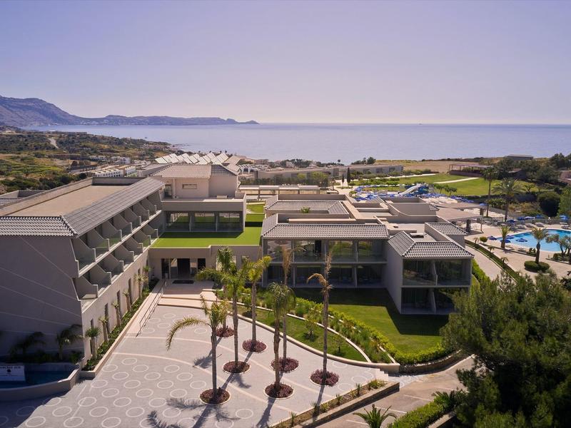 Complejo hotelero con piscina y palmeras junto al mar con montañas al fondo.