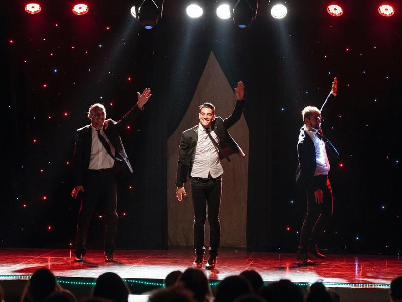 Tres hombres bailan sincronizados en un escenario con luces rojas de fondo.