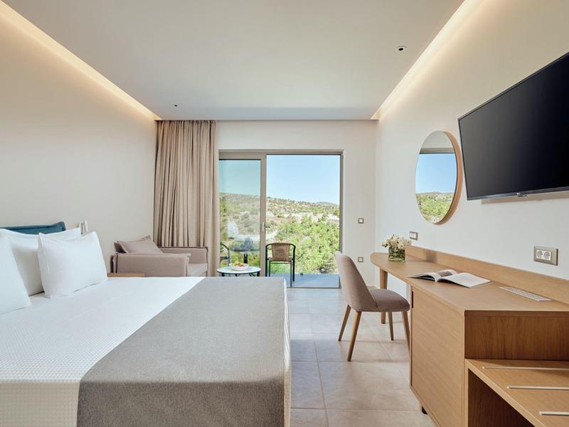 Habitación de hotel luminosa con cama doble, mobiliario de madera, TV y balcón con vista a colinas verdes.