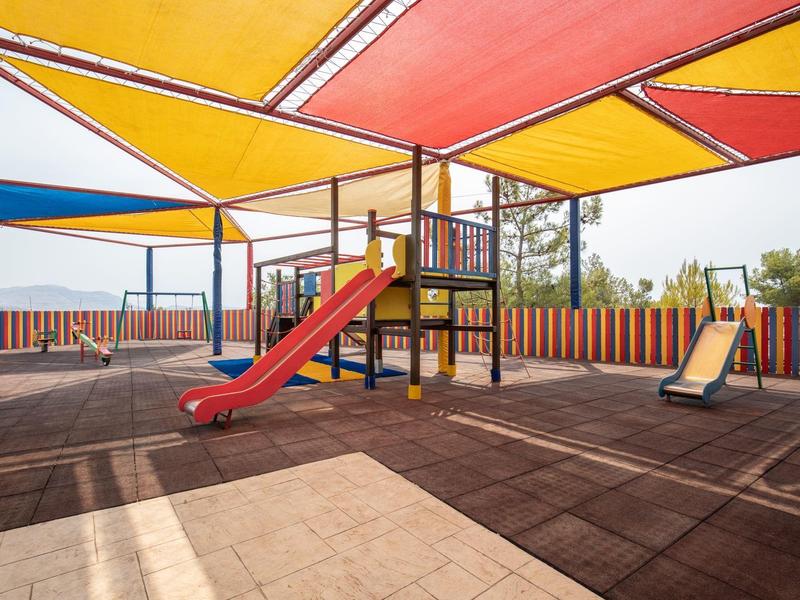 Área de juegos con toboganes bajo toldos rojos y naranjas, rodeada de una valla de madera colorida.