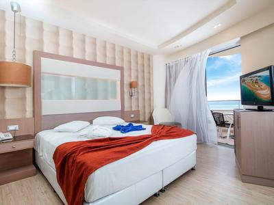 Bright hotel room with double bed, red blanket, TV, and balcony with sea view.