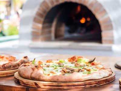 Świeżo upieczona pizza na drewnianej desce z piecem opalanym drewnem w tle.