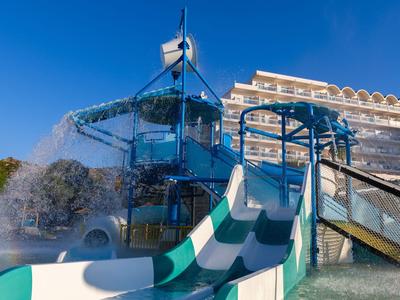 Wasserrutsche und Wasserspielplatz vor einem Hotel unter klarem blauem Himmel.