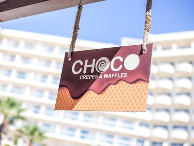 Schild mit Aufschrift 'CH@CO Crepes & Waffles' vor einem Hotel mit blauen Himmel im Hintergrund.