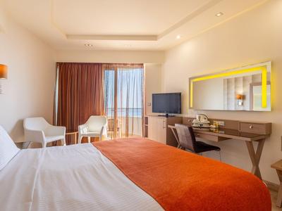 Helles Hotelzimmer mit Doppelbett, orangener Bettdecke, Schreibtisch und Sitzbereich mit zwei Stühlen.