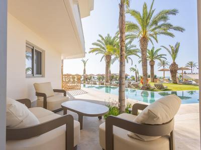 Hotelterrasse mit bequemen Sesseln und Pool umgeben von Palmen bei Sonnenschein.