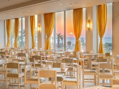 Helles Restaurant mit weißen Stühlen und großen Fenstern mit Blick auf Sonnenuntergang und Meer.