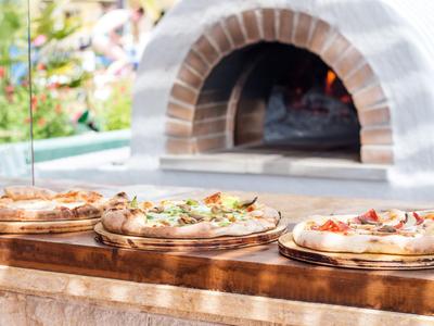 Drei belegte Pizzen vor einem Holzofen, bereit zum Servieren im Freien.