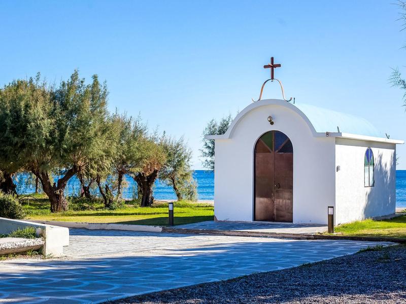 Small white chapel with wooden door and cross by the beach with olive trees in sunlight.