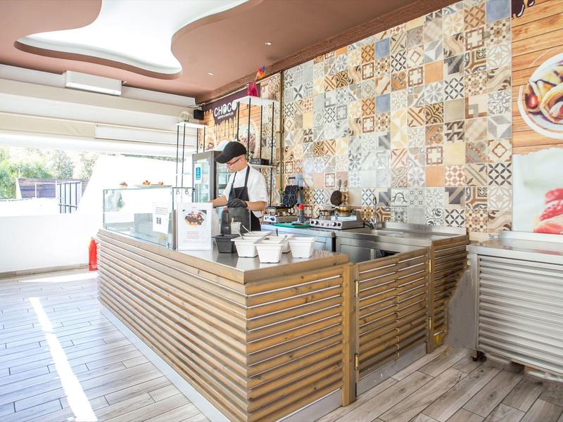 Modern kiosk with wooden counter and colorful tiled wall, open to a sunny outdoor area.