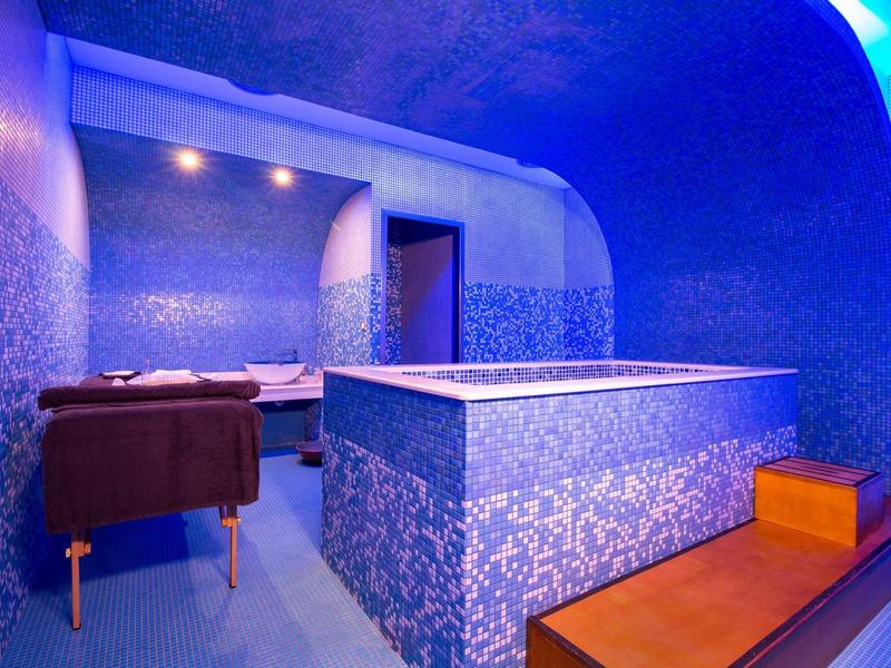 Bright spa with blue tiled bathtub, lounge chairs, and ambient lighting.