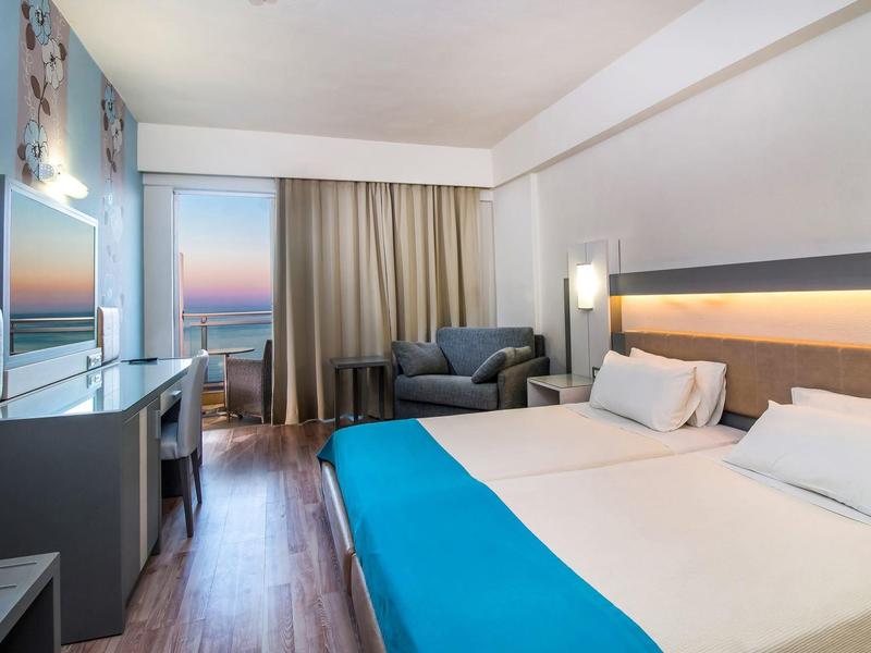 Modern hotel room with double bed, sofa, TV, and window showing sunset view.