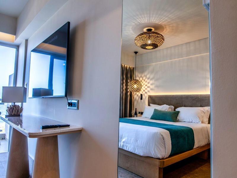Mirror reflects modern hotel room with bed, green accent, and ceiling light.