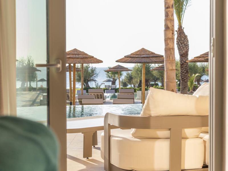 View from indoors to a terrace with lounge chairs, umbrellas, and palm trees.