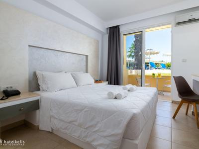 Světle zařízený hotelový pokoj s manželskou postelí, balkonem a výhledem na bazén a zahradu.