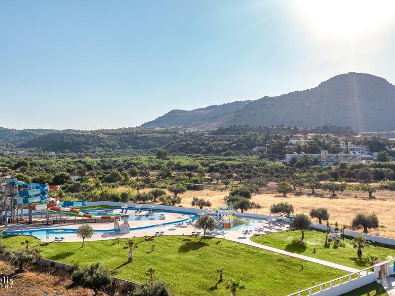 Prostorný hotelový komplex s bazénem, obklopený zelenými loukami a horami v pozadí za slunečného počasí.