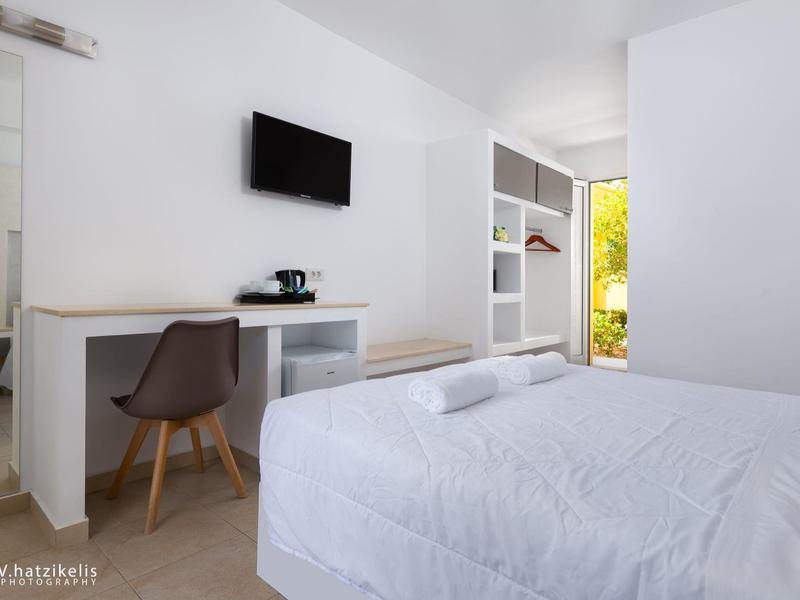 Světý hotelový pokoj s bílou postelí, psacím stolem, židlí a televizí na zdi.