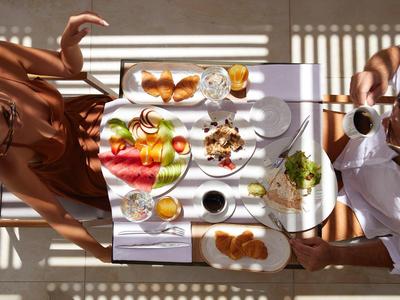 Zwei Personen beim Frühstück an einem weißen Tisch mit Kaffee, Croissants, Obst und Eiern im Sonnenlicht.