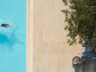 Ein Schwimmer im blauen Pool neben einer hellen Steinplatte und Schatten von Bäumen.