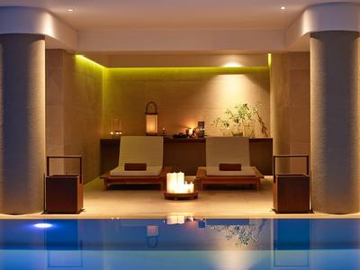 Moderner Wellnessbereich mit beleuchtetem Pool, zwei Liegen und warmem Licht an der Wand.
