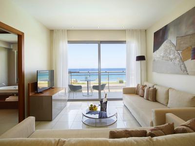 Helles Wohnzimmer mit beige Sofa, Glastisch, großem Fenster und Blick auf das Meer und Balkon.