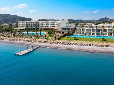 Großes Hotel mit Pool, Liegestühlen und kleinem Steg am Ufer eines ruhigen blauen Meeres.