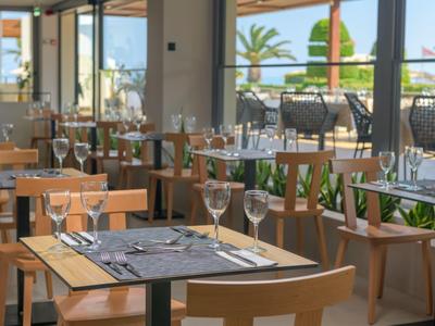 Moderner Innenraum eines Restaurants mit Holztischen, Stühlen und großen Fenstern zum Außenbereich.