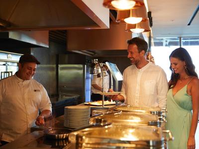 Ein Paar steht an einem Buffet und wählt Essen aus, während ein Koch lächelnd daneben steht.