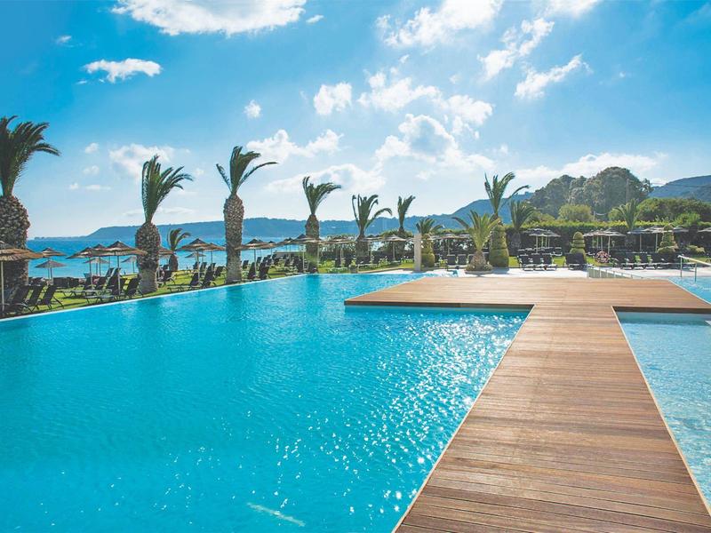 Großer blauer Pool mit Palmen, Holzsteg und blauem Himmel mit weißen Wolken im Hintergrund.