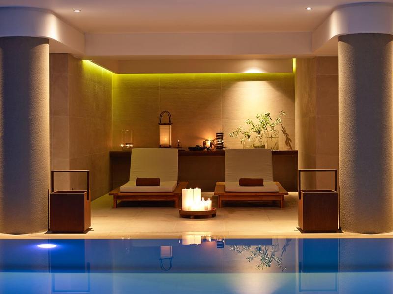 Moderner Wellnessbereich mit beleuchtetem Pool, zwei Liegen und warmem Licht an der Wand.