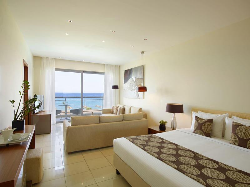 Helles Hotelzimmer mit großem Bett, Sofa, Fernseher und Balkon mit Meerblick.