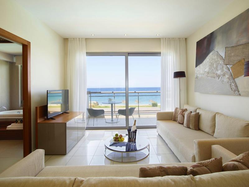 Helles Wohnzimmer mit beige Sofa, Glastisch, großem Fenster und Blick auf das Meer und Balkon.