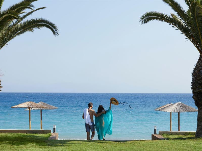Paar geht von einer Rasenfläche zwischen zwei Palmen zum Strand mit Sonnenschirmen.