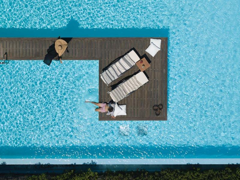 Überblick aus der Vogelperspektive eines hellblauen Pools mit Sonnenliegen auf einer Mitteinsel.