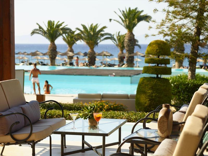 Sitzbereich mit Sofas und Tisch vor einem Pool mit Palmen und Meer im Hintergrund.