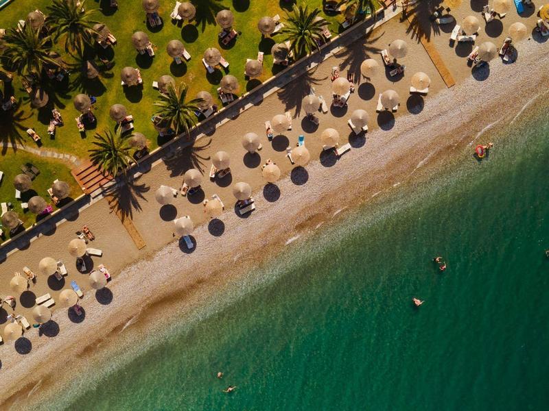 Luftaufnahme eines Strandes mit Sonnenschirmen, Palmen und klarem, grünem Wasser.