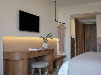 Hotelzimmer mit Fernseher, Holzmöbeln, Sitzhocker und weißer Vase auf Schreibtisch.