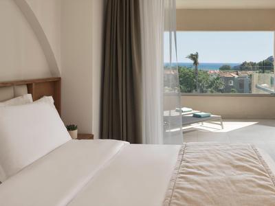 Camera d'hotel luminosa con letto bianco, tenda grigia e terrazza con vista mare.