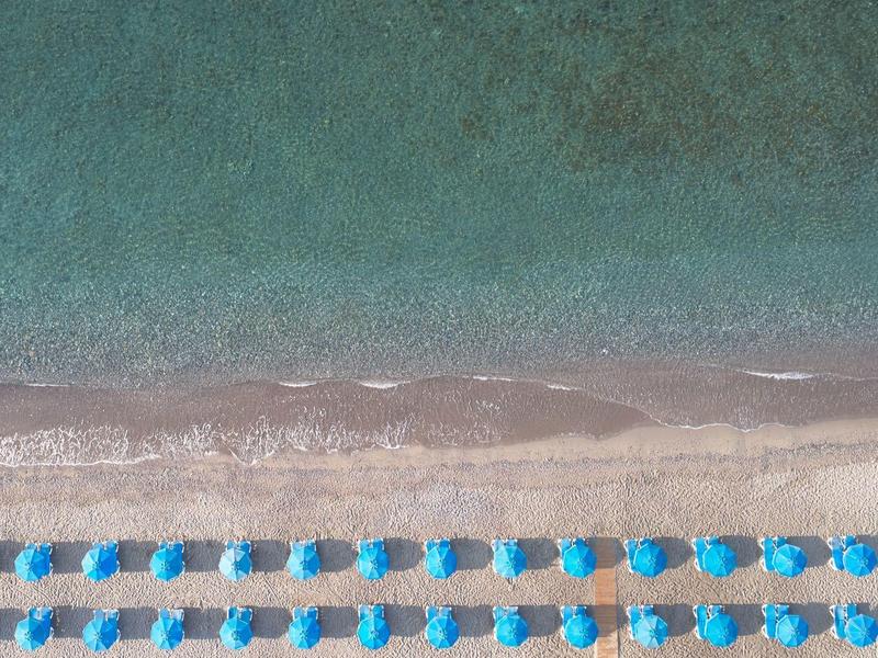 Vista aerea di una spiaggia con file di ombrelloni blu sull'acqua cristallina.