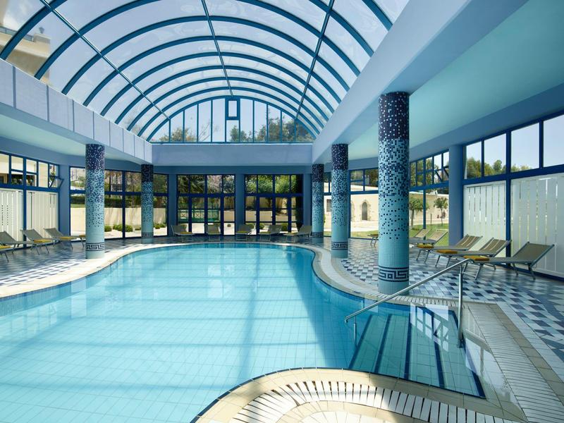 Grande piscina coperta con cupola di vetro, colonne e lettini ai lati.