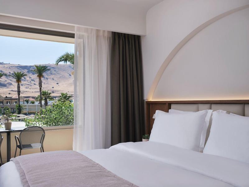 Camera d'albergo luminosa con letto matrimoniale e balcone con vista su palme e montagne.