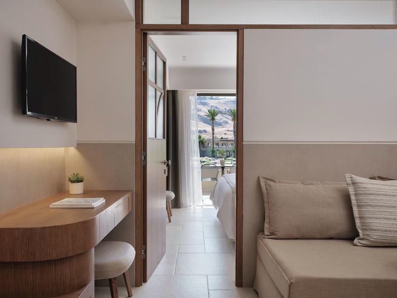 Zona soggiorno luminosa in hotel con divano, TV e vista sul balcone con palme.